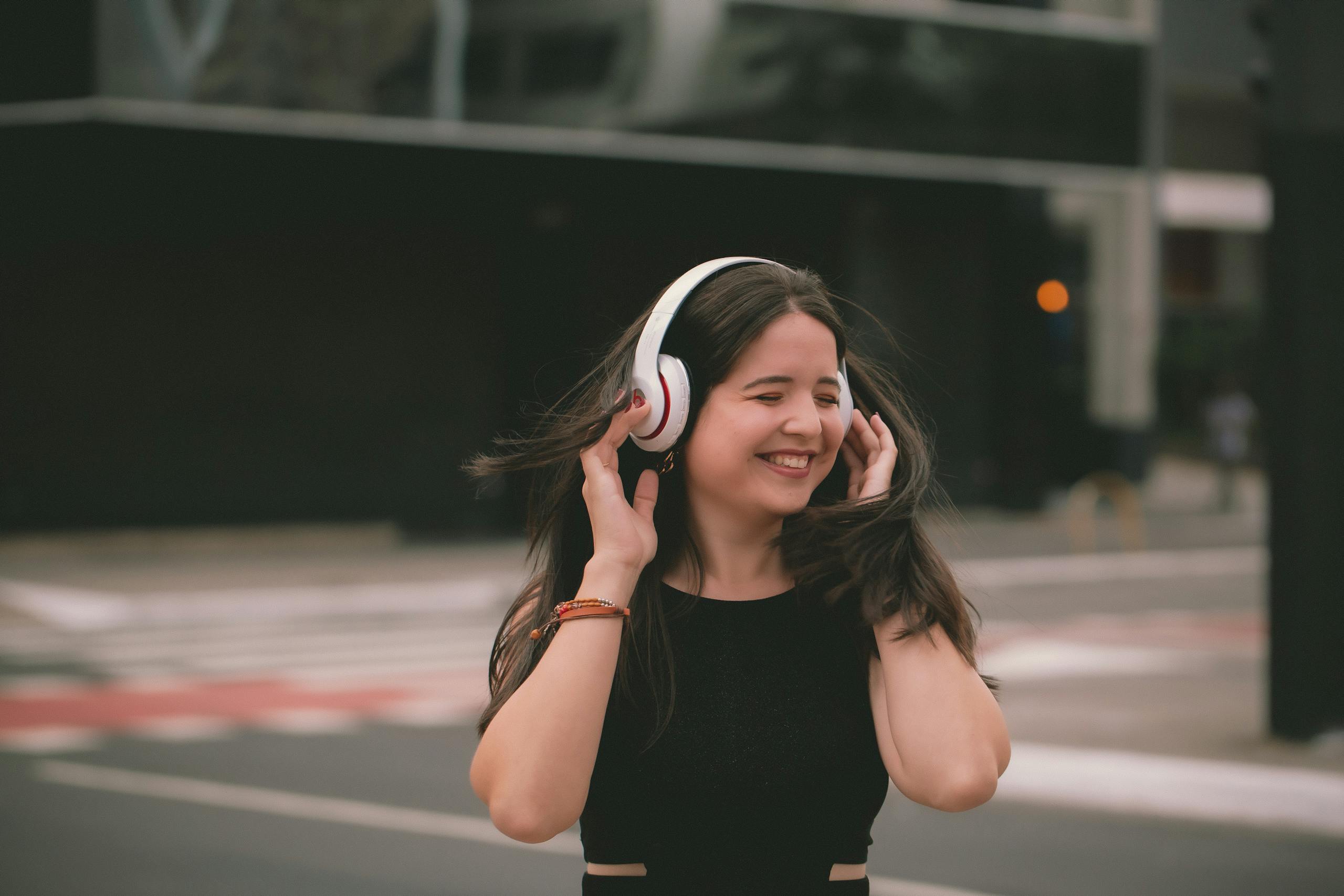A smiling woman listens to music wearing wireless headphones in an urban setting, expressing happiness.